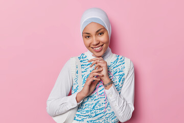 Photo of charming pleased beautiful Muslim woman smiles pleasantly shows white teeth touches chin gently looks directly at camera wears traditional hijab and vest poses against pink background
