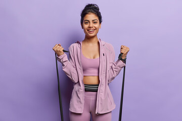 Indoor photo of young pretty smiling happy European lady standing isolated on purple background wearing tracksuit doing physical exercises using expander as part of daily routine to keep fit