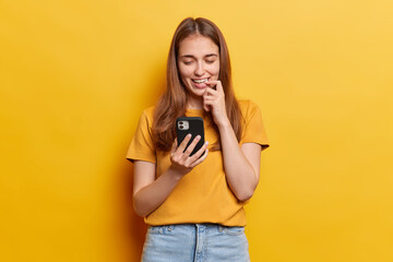 Professional indoor shot of attractive young european sming woman with long straight loose hair isolated on yellow background looking delighted reading message with good news on her smartphone