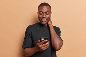 Pleased dark skinned man keeps hand on neck smiles pleasantly holds mobile phone watching video using entertaining mobile application dressed in casual black t shirt isolated over brown background.