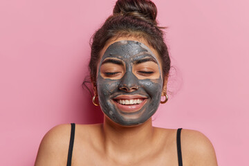 Studio shot of young glad european pretty girl applying natural beauty clay mask to keep her skin in good conditions smiling happily with closed eyes keeping her hair in bun. Beauty skincare concept