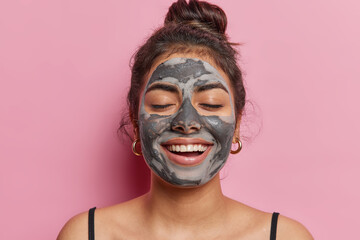 Skincare and wellness concept. Horizontal indoor close up of young pretty pleased woman smilling broadly and happily with closed eyes applying natural beauty clay mask to make her wrinkless less