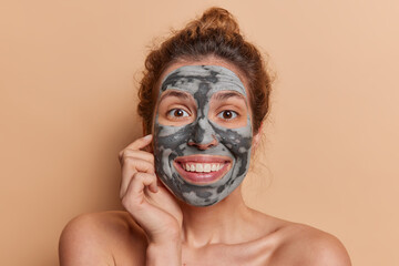 Headshot of cheerful young pretty European woman applies clay mask for achieving naturally beautiful and rejuvenated look smiles broadly stands bare shouldered against brown studio background