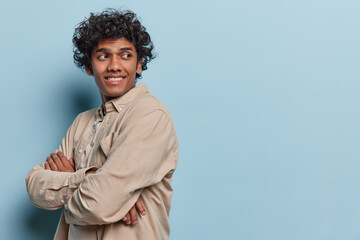 Obraz premium Sideways shot of curly haired Hindu teenage man keeps arms folded looks behind smiles gladfully notices something pleasant isolated over blue background copy space for your advertising content