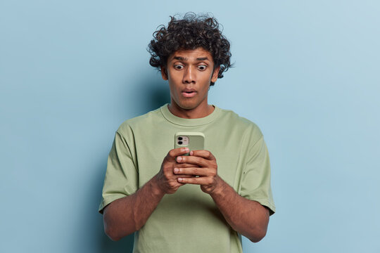 Waist up shot of stunned curly haired Hindu man being deeply surprised stares at smartphone display reads shocking news on website notices something horrible wears green t shirt isolated on blue wall