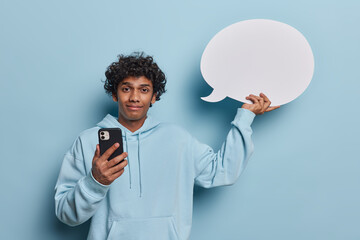Waist up shot of pleased youngHindu man with curly hair holds smartphone and communication bubble...