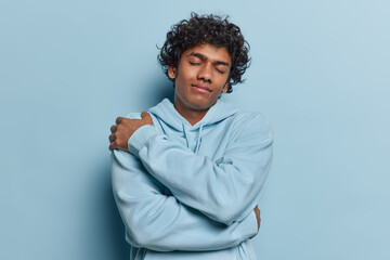 Waist up shot of pleased Hindu man with curly hair embraces himself keeps eyes closed dreams about something dressed in comfortable sweatshirt isolated over blue background. Love yourself concept - obrazy, fototapety, plakaty