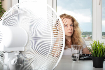 Woman blowing a fan to prevent sweating and stay comfortable in the heat in the office, Preventing...