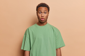 People emotions concept. Professional studio shot of young surprised African male with curly dark hair wearing casual green tshirt standing in centre isolated on beige background keeping hands down