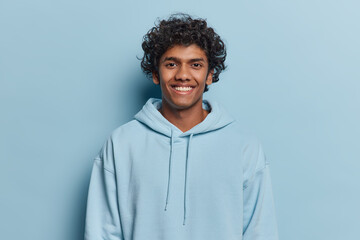 Portrait of handsome curly haired young Hindu man smiles pleasantly dressed in casual hoodie expresses positive emotions poses against blue studio background. Monochrome shot. Youth concept. - 624029232