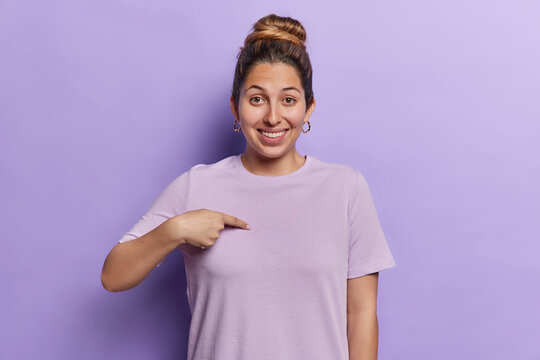 Waist up shot of cheerful beautiful European woman points at herself asks who me smiles pleasantly glad to be chosen dressed in casual t shirt isolated over purple background. Do you mean me