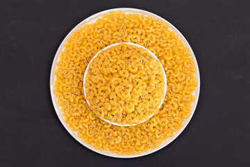 Uncooked Chifferi Rigati Pasta in White Plate on Black Background. Fat and Unhealthy Food. Scattered Classic Dry Macaroni. Italian Culture and Cuisine. Raw Pasta