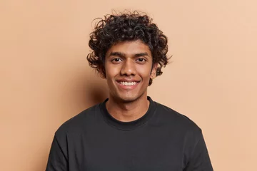 Fotobehang Portrait of handsome cheerful man with curly hair smiles toothily poses happy against brown background dressed casually isolated over brown background. Positive human emotions and ethnicity concept © Wayhome Studio