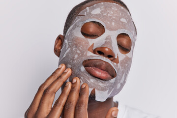 Close up shot of calm dark skinned man applies moisturising sheet mask on face keeps eyes closed...