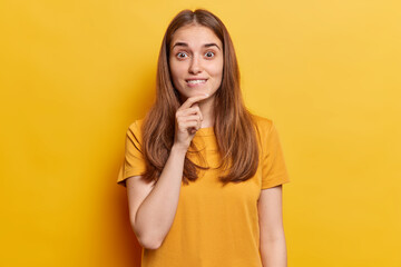 Beautiful glad young european woman with long loose hair looks straight with happy expression thinks about pleasant things keeping hand under chin wears casual tshirt isolated on yellow background