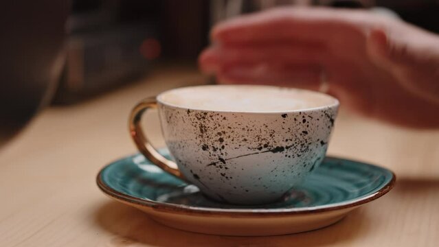 Female hand of the barista puts the finished beautiful cappuccino coffee in a cup on a saucer on the wooden table. Slow motion closeup shot with bokeh