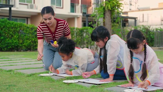 Group of diverse elementary students and female teacher learn art and drawing poster color outdoors at lawn. Children use time to activity creativity during school lesson in park at elementary school.