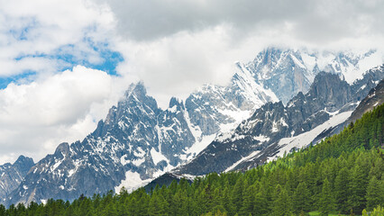 Fototapeta premium The famous Monte Bianco's peak (White Mountain) covered by dark clouds, in Aosta Valley, Italy. Green pine forest in the foreground. Clouds on the background.
