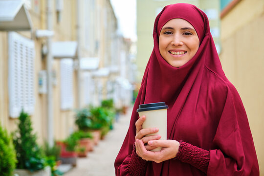 Portrait of muslim young woman in hijab smiling holding a coffee cup in the street and looking at camera.