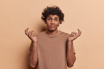 Difficult choice concept. Hesitant Hindu man with curly hair shrugging shoulders snd spreading arms says who knows canot find clue dressed in casual t shirt isolated over brown studio background.