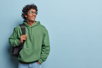 Indoor waist up of young smiling happy Hindu man standing on left isolated on blue background wearing casual green hoodie and jeans with bag on left shoulder looking at blank space for your promotion
