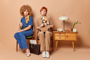 Two thoughtful women dressed in vintage attire pose in cozy room adorned with retro furniture wait in queue near cabinet dont look at each other sit on chairs isolated over brown background.