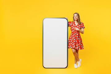 Full body young woman wear red dress casual clothes point index finger on big huge blank screen mobile cell phone smartphone with workspace copy space mockup area isolated on plain yellow background.