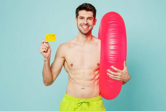 Young fun man wear green shorts swimsuit relax near hotel pool hold inflatable rubber ring hold credit bank card isolated on plain light blue cyan background. Summer vacation sea rest sun tan concept.