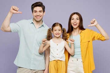 Young smiling happy parents mom dad with child kid daughter girl 6 years old wear blue yellow casual clothes point thumb fingers on themselves isolated on plain purple background. Family day concept.