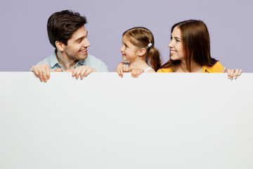 Young amazed parents mom dad with child kid daughter girl 6 years old wearing blue yellow casual clothes hold big white empty poster billboard isolated on plain purple background. Family day concept.