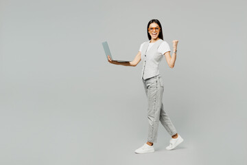 Full body young latin IT woman she wear white t-shirt gray vest glasses hold use work on laptop pc computer do winner gesture walk isolated on plain grey background studio portrait. Lifestyle concept.