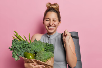 Overjoyed European woman with hair bun keeps eyes closed clenches fist keeps to healthy diet carries vegetables in paper bag uses fitness mat for pilates exercises isolated over pink background