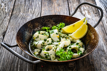 Fried calamari rings with garlic, lemon and parsley in pan on wooden table
