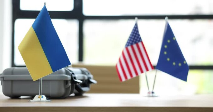 Small flags of USA and European Union on board table near small flag of Ukraine. Flags hide suitcase with secret contents. Foreign financial aid to Ukraine in war