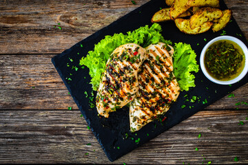 Grilled chicken breasts with Argentinian chimichurri sauce,  baked potatoes and fresh vegetables on wooden table
