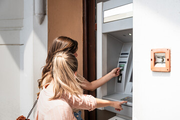 Two beautiful tourists female withdrawing the money from a bank card using ATM machine at the city...