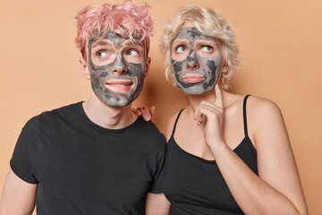 Studio shot of thoughtful young European male and female doing cosmetology procedures applying natural clay mask on faces as part of daily routine to reduce pimples and improve condition of skin