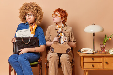 Photo of two businesswomen occupy chairs in acozy room adorned with retro furniture pose with briefcase full of papers look puzzled wear spectacles and elegant clothing isolated over brown background