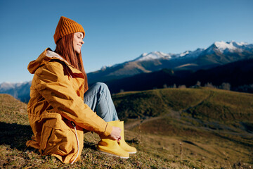 Woman sitting on a hill on the grass rest smile with teeth looking at the mountains in the snow in the autumn in a yellow raincoat and jeans happy sunset hiking trip, freedom lifestyle 