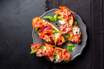 A dark grey plate with with open sandwiches, snack. Toastet bread, griddled peach, parma ham or...