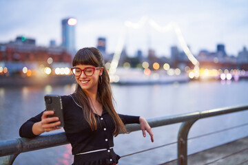 portrait beautiful young adult woman looking at a cell phone and smiling at sunset in the city