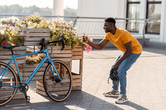 Smiling attractive young African man holding mobile phone, taking photo of bicycle on street