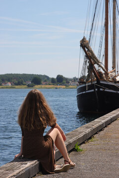 Attraktive, junge Frau mit langen Beinen und  braunen Haaren im Sommerkleid sitzt am Kai von Svendborg mit Segelschiff.
