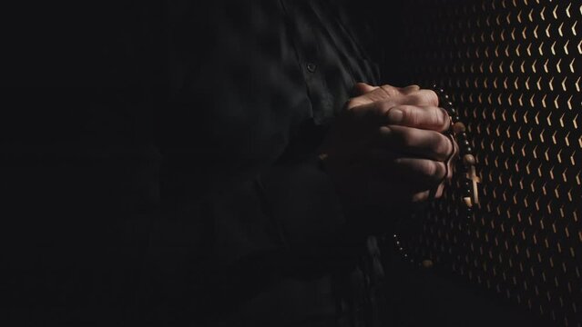 Close-up of unrecognizable Catholic priest holding rosary beads in hands carefully listening to penitent in confessional