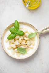 Gnocchi with cheese sauce and basil in a serving pan, top view on a light-beige stone background, vertical shot
