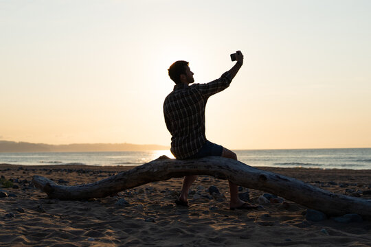 Silhouette of a man sitting on a driftwood trunk holding a cellphone taking a selfie outside during sunset on the beach. Black light photo.