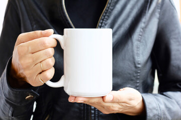 Girl is holding white mug in hands with black leather jacket. Blank 15 oz ceramic cup