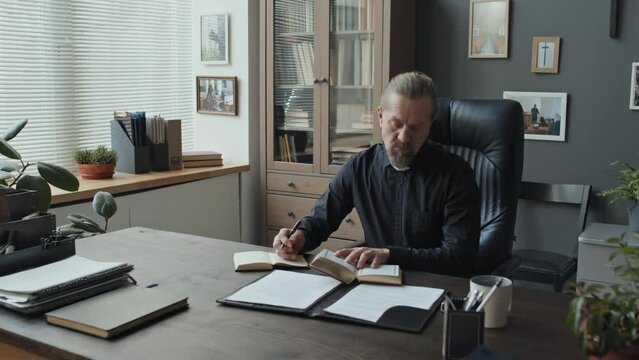 Modern mature Catholic priest sitting desk in his office reading Bible and making notes in notebook