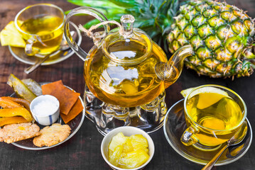 Glass tea pot with pineapple tea and homeade jam,cups and crystalized fruits on dark background