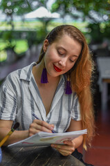 Beautiful blonde woman artist and illustrator thinking about a concept of her drawing, sitting by the table with drawings, brushes and paint